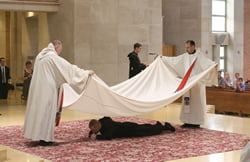 Photo by JD Benning Benedictine Father Marion Charboneau (left) and Prior James Albers cover Brother Leven with a funeral pall to symbolize his death to the world. Brother Leven emerges from beneath the pall, symbolically resurrecting to new life as a monk.