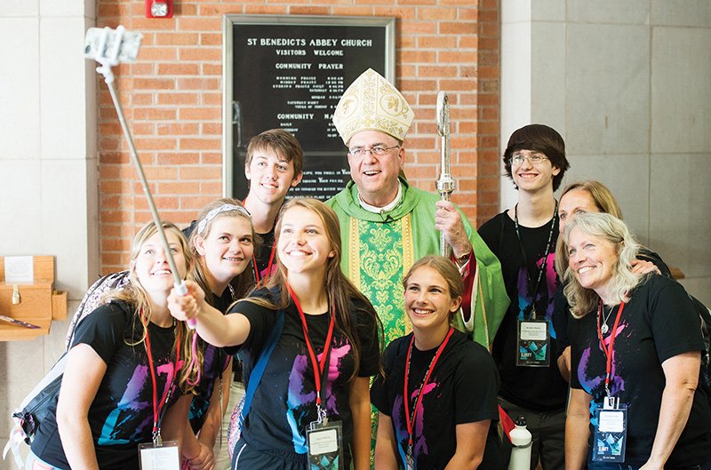 Some archdiocesan teens, part of the Northeast Kansas Rural Youth Council, pose with Archbishop Joseph F. Naumann for a picture with a selfie stick after the Life Teen Leadership Conference Mass at St. Benedict’s Abbey in Atchison on June 25.  The teens are, from left, Marley Wareham, St. Dominic Parish, Holton; Sami Fischer, St. Stanislaus Parish, Rossville; Wade Minihan, St. Columbkille Parish, Blaine; Taylor Bittner, St. Stanislaus; Nikita Rogers, St. Francis Xavier Parish, Burlington; and Braden Myers, St. Francis Xavier. Also pictured are, second from right, Angie Bittner, archdiocesan rural youth ministry outreach coordinator, and Michelle Giesy, St. Patrick Parish, Osage City.  By Jessica Langdon